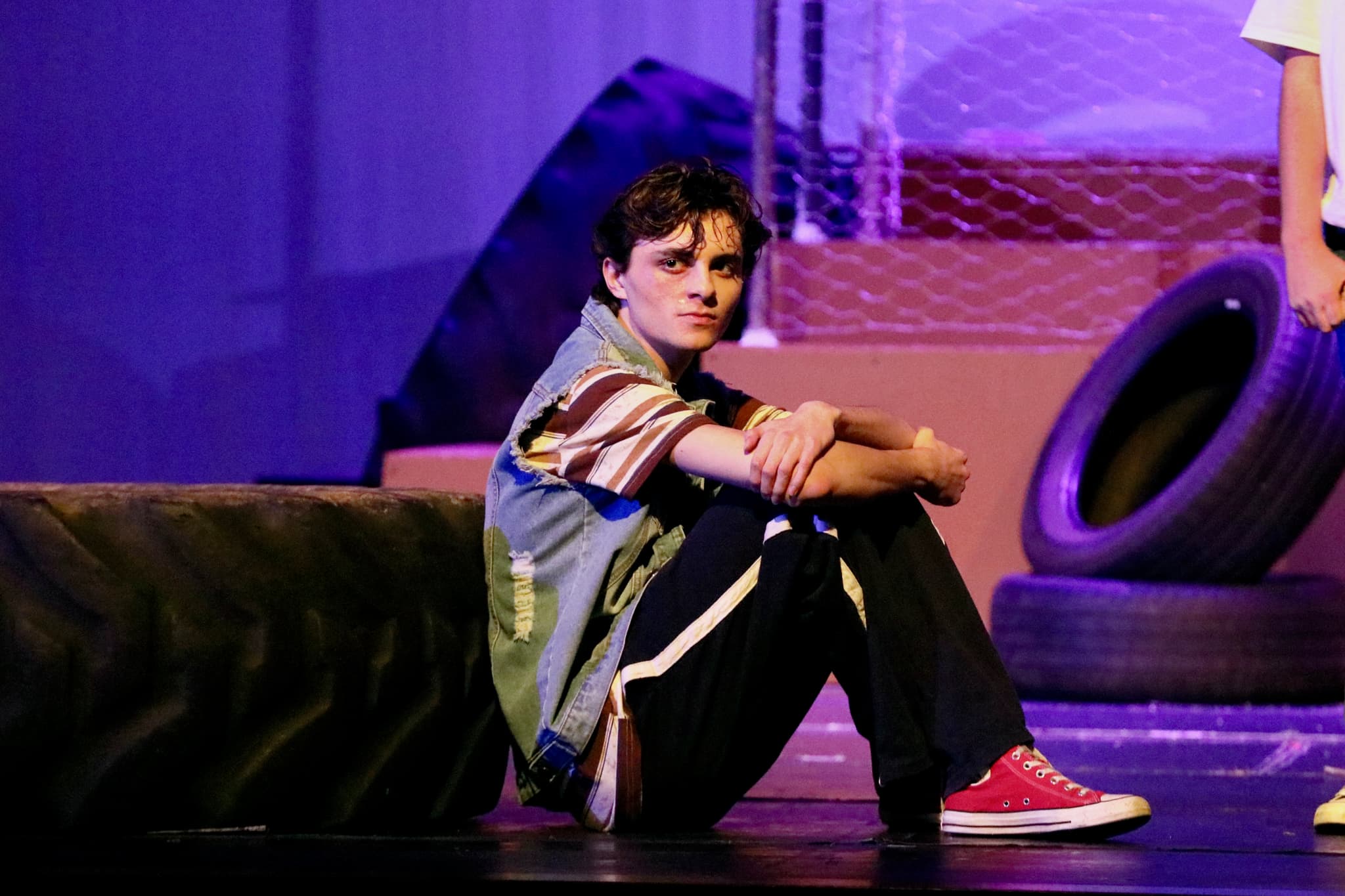 Young actor sits on stage floor between tires under blue light