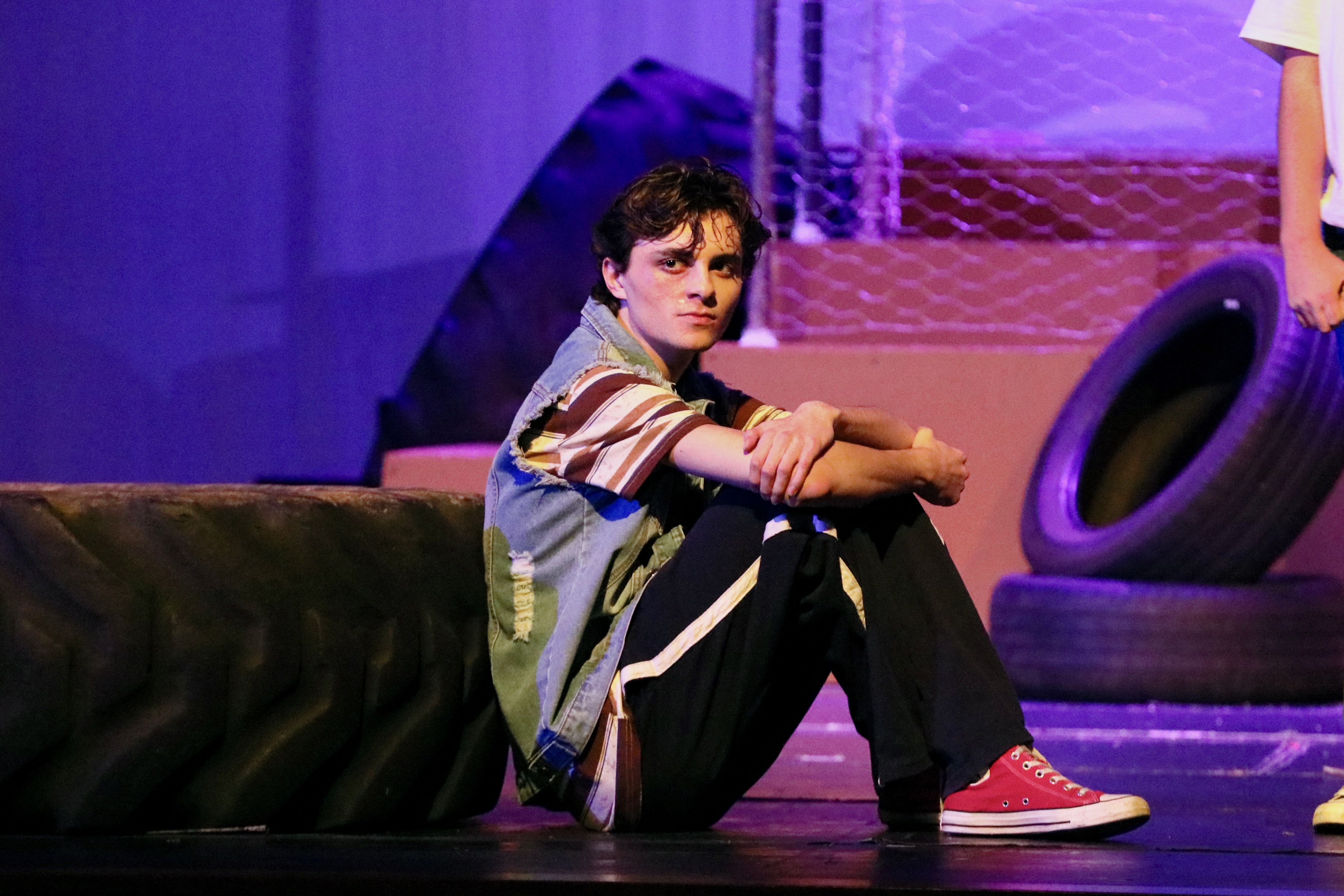 Young actor sits on stage floor between tires under blue light